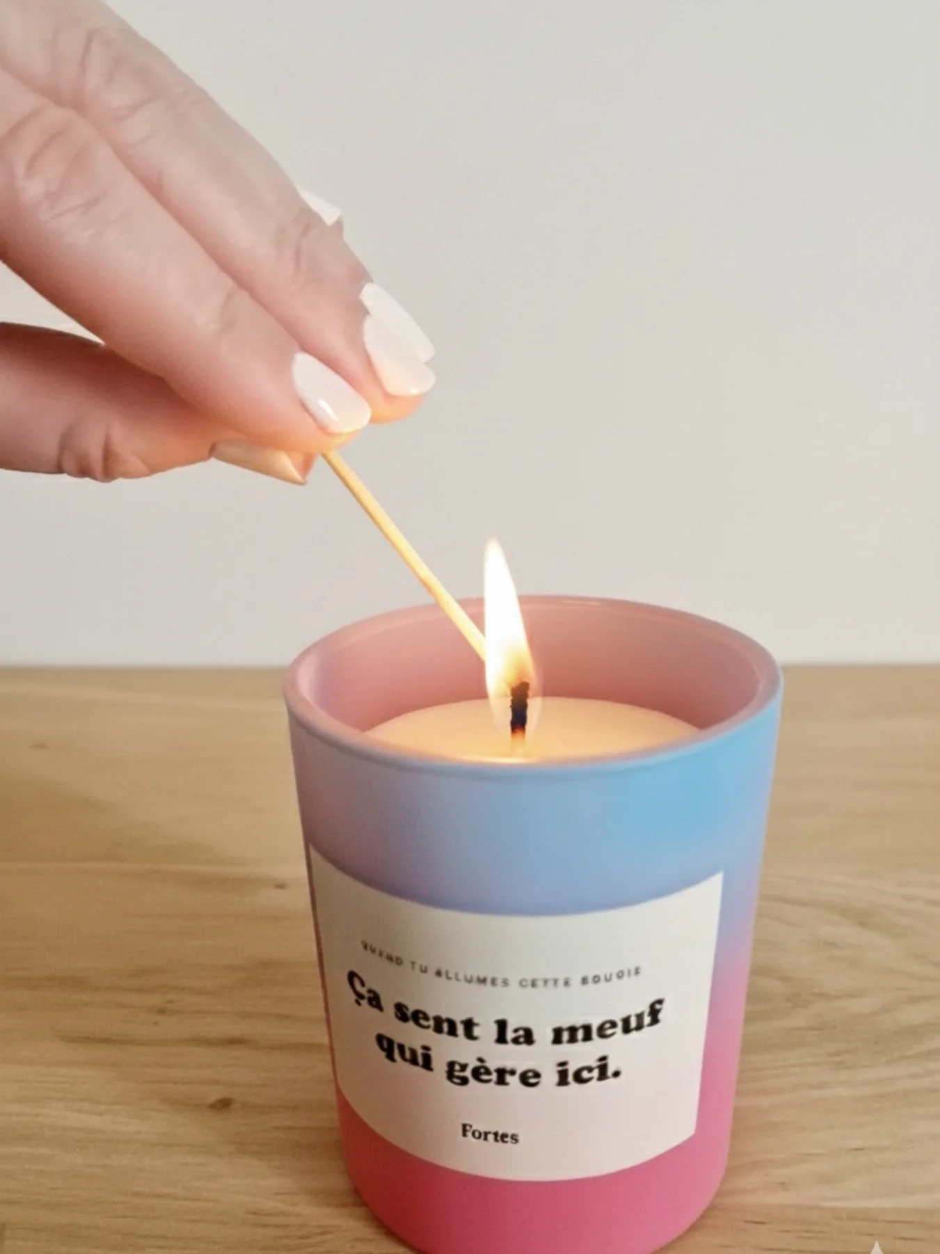 Main avec manucure blanche allumant une bougie parfumée colorée sur une table en bois