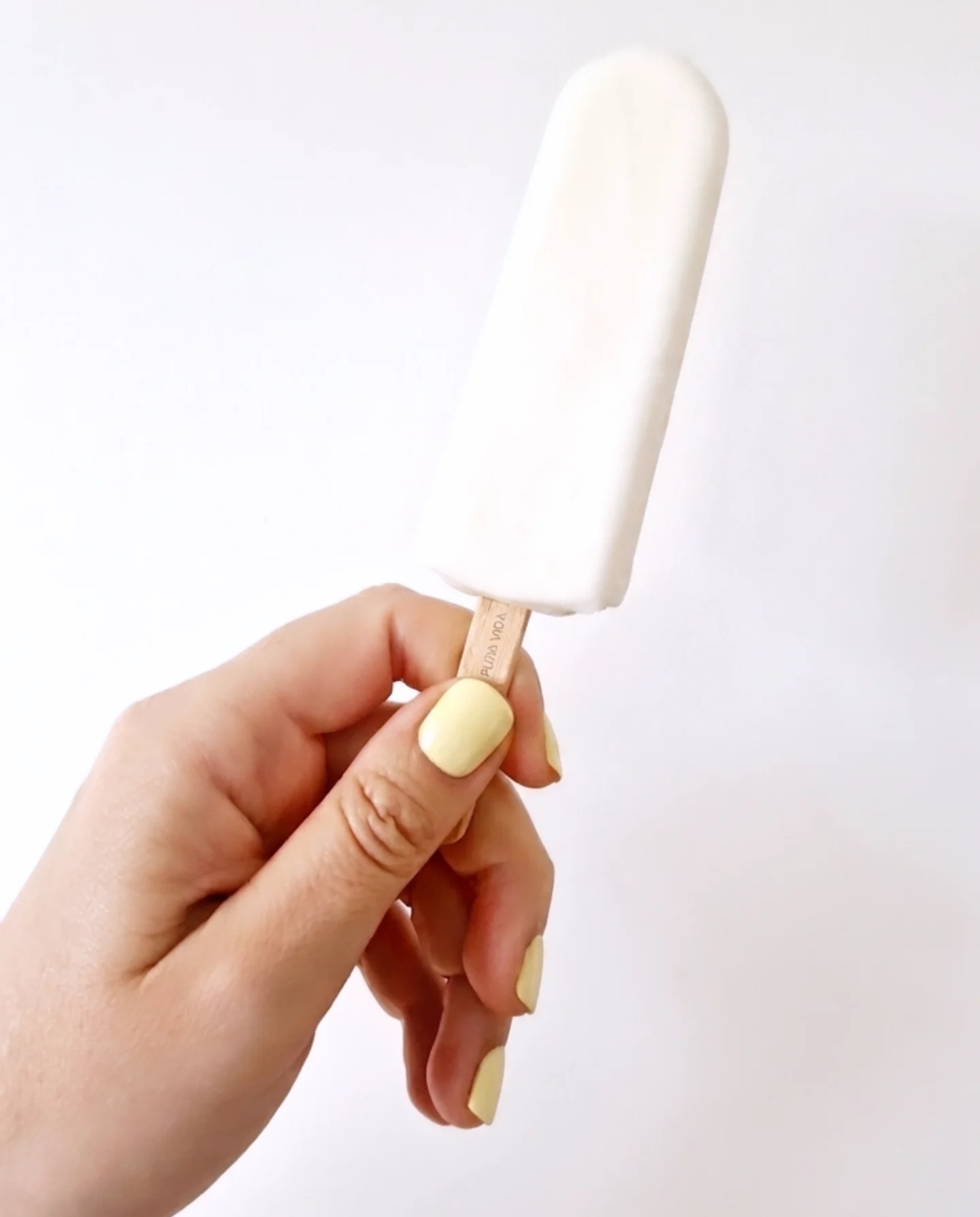 Main avec manucure jaune pastel tenant une glace blanche sur fond neutre